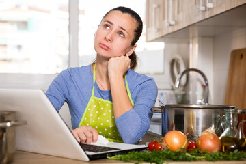 Frustrated brunette woman looking for recipe for soup on Internet at kitchen