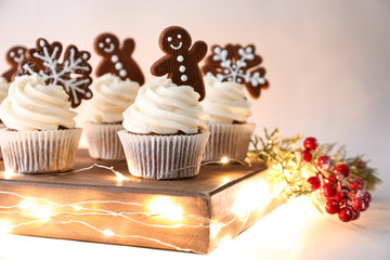 Wooden board of tasty cupcakes with gingerbread cookies and Christmas lights on beige background