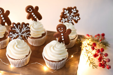 Wooden board of tasty cupcakes with gingerbread cookies and Christmas lights on beige background