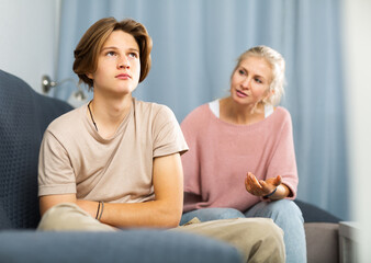 Portrait of upset and offended teenager sitting on sofa while mother soothing him. Mood swings and...