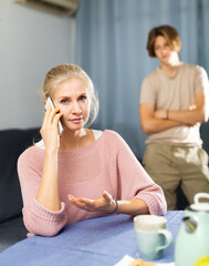 Woman having serious phone conversation while sitting at table at home against background of her upset teenage son