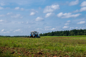 Tractor working in the field, plowing the land, preparing for sowing. View of the field and tractor in the distance against a blue sky with clouds