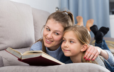 Portrait of happy young woman and her pre-teen daughter reading book at home