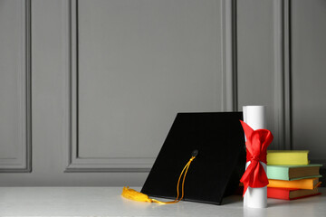 Graduation hat, books and diploma on white table near grey wall, space for text