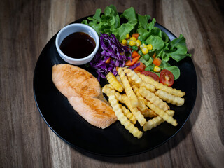 Grilled salmon steak with vegetable and french fries on plate, on wood table, top view.