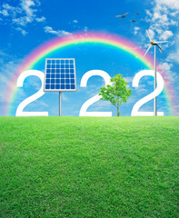 2022 white text with solar cell, wind turbine and growing tree on green grass field over rainbow, birds and blue sky with white clouds, Happy new year 2022 ecological cover concept