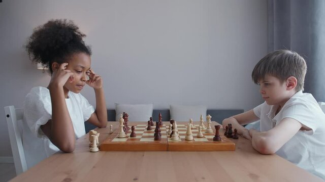 Children in self-isolation, an African girl and a Caucasian boy play chess sitting in the living room, entertainment during quarantine, the thought process.