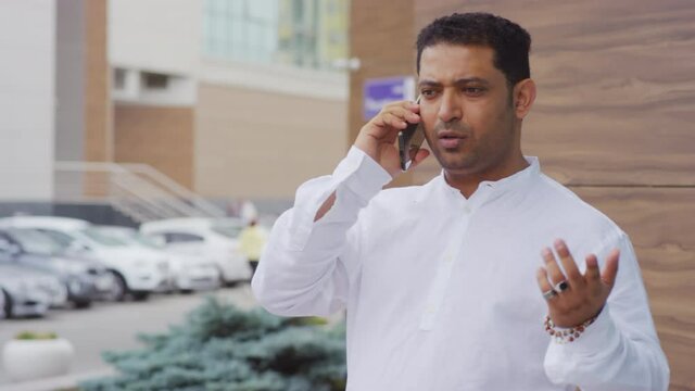 Chest-up of young Middle Eastern man wearing white shirt talking on cellphone, standing by wall of building outside