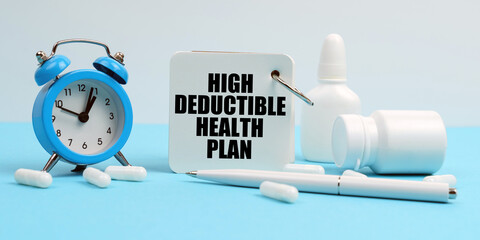 On a blue surface lie pills, an alarm clock and a notebook with the inscription - High Deductible Health Plan