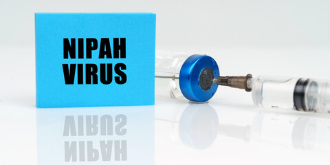 On a white reflective surface are a syringe, an injection and a blue plaque that says - Nipah virus