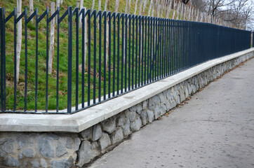 fencing made of metal bars painted gray to black. the web is joined at the top diagonally downwards. The fence is elegantly transparent with a stone wall foundation