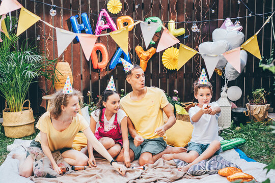 Little Boy Closes Ears While Parents With Sister Use Party Popper Sitting On Plaid At Birthday Celebration In Decorated Cottage Yard In Summer