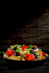 Salad with tomatoes, peppers, tuna olives and pasta. Fresh salad in a black plate on a dark background. Copy space and free space for text near food.