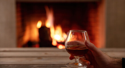 Man holding a brandy glass on burning fireplace background. Drink alcohol and relax, winter time.