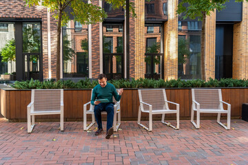 Fototapeta na wymiar Millennial man with happy face checking phone sitting on one of four chairs in courtyard of modern brick residential complex. Portrait of excited man holding smart phone at street outdoors.