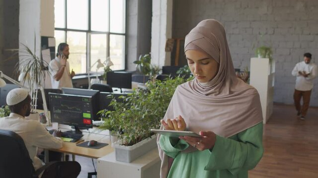 Waist-up of portrait of hazel-eyed Caucasian woman wearing hijab, using tablet computer, standing in office at daytime, then looking up and smiling on camera