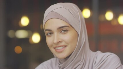 Close-up of young Caucasian hazel-eyed Muslim woman wearing hijab looking and smiling on camera, blurred lights on background
