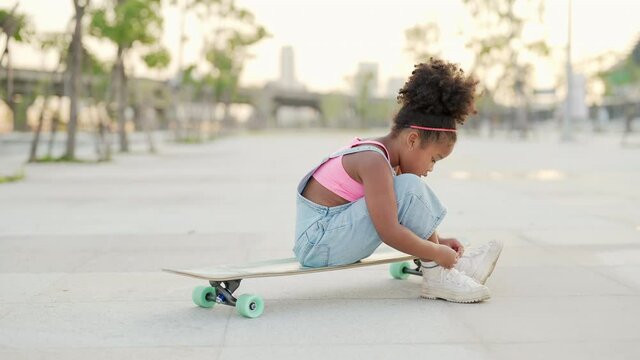 4K African child girl kid tying sneaker shoes while skateboarding at the park. Cute preschool girl enjoy and having fun outdoor lifestyle practicing extreme sport longboard skating on summer vacation.