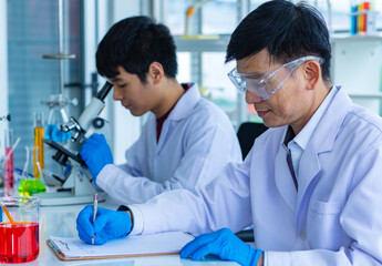 Portrait shot of Asian young male scientist in white lab coat and rubber gloves sitting smiling using microscope in laboratory while colleague wearing safety goggles writing result on paper clipboard