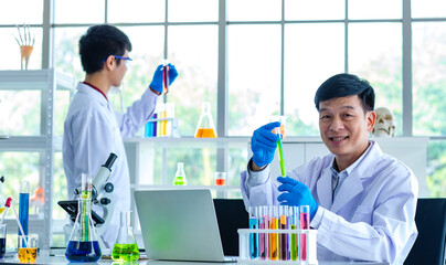 Portrait shot of Asian professional mature male scientist in white lab coat and rubber gloves sitting look at camera smiling holding blue liquid sample in test tube while collegaue working behind