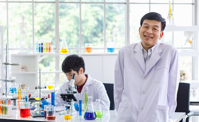 Portrait shot of Asian professional successful mature male scientist in white lab coat rubber gloves standing crossed arms smiling look at camera in laboratory while young colleague working behind