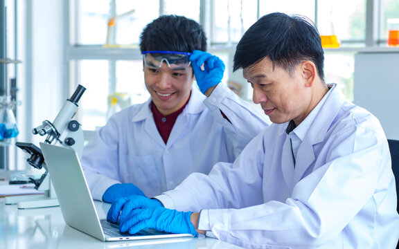 Asian professional mature male scientist teacher in white lab coat and rubber gloves sit smiling teaching science student using microscope and record analysis data in laptop computer in laboratory