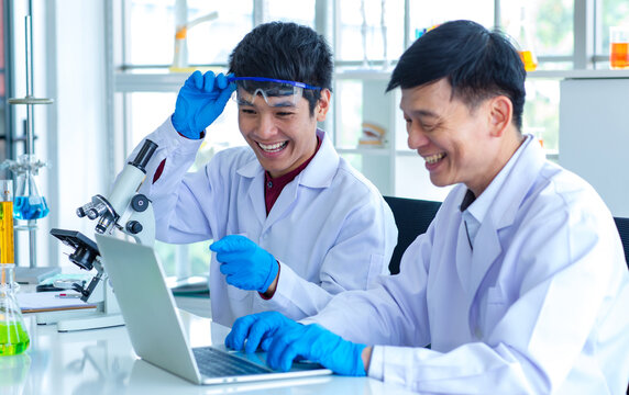 Asian professional mature male scientist teacher in white lab coat and rubber gloves sit smiling teaching science student using microscope and record analysis data in laptop computer in laboratory