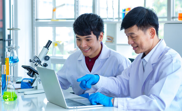 Asian professional mature male scientist teacher in white lab coat and rubber gloves sit smiling teaching science student using microscope and record analysis data in laptop computer in laboratory - Powered by Adobe
