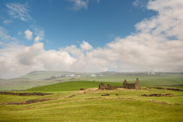 Obraz premium Beautiful rural landscape scene. Small stone abandoned house on a green grass hill, mountains and houses in the background. Cloudy sky. West of Ireland. Calm and peaceful mood.