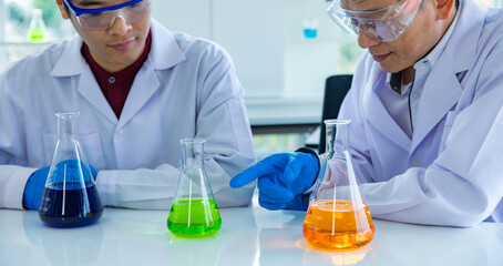 Two Asian male scientists in white lab coat safety goggles rubber gloves working discussing talking together about colorful liquid samples in glass flasks in scientific experiment on laboratory table