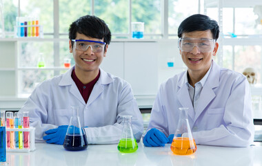 Two Asian male scientists in white lab coat safety goggles rubber gloves working discussing talking together about colorful liquid samples in glass flasks in scientific experiment on laboratory table