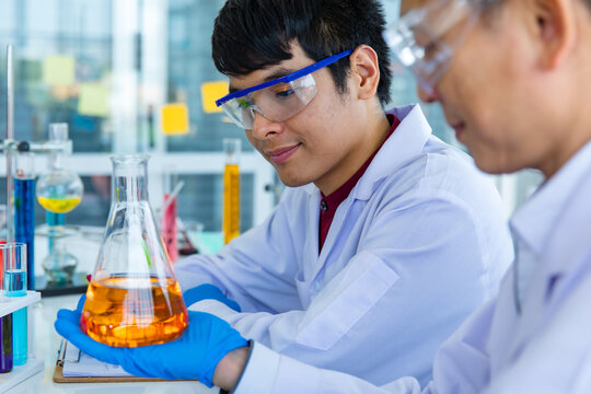 Portrait closeup shot of Asian mature male scientist teacher in white lab coat safety goggles and rubber gloves using microscope smiling look at camera with university student in blurred background