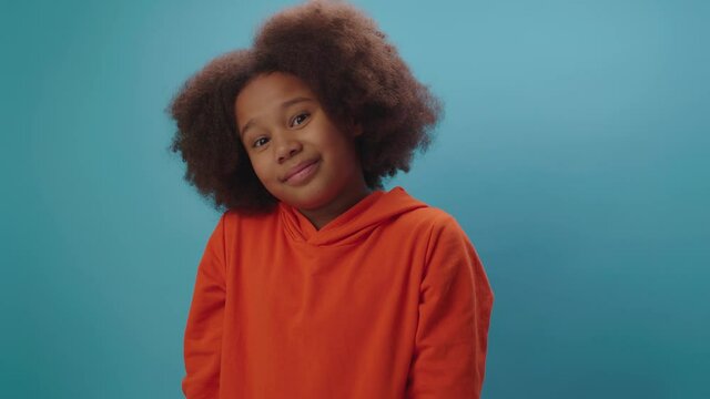 Young African American girl showing the emotion 'I don't know'. Kid shrugs doubtful standing on blue background. Hesitating child doesn't know.