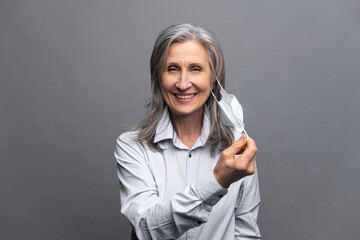 Happy senior woman in formal wear taking off her medical mask isolated on gray. Close-up portrait of female office employee protecting herself from viral disease. Recommends measures concept