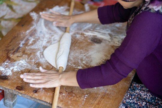 Female Hands Of Baker Rolls Dough In Flour On Wooden Board On Table With A Rolling Pin, In Bakery Shop, Woman Making Pizza Using Traditional Recipe At Home.