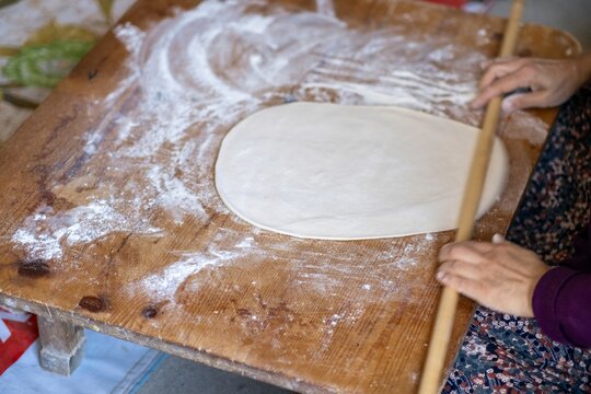 Female Hands Of Baker Rolls Dough In Flour On Wooden Board On Table With A Rolling Pin, In Bakery Shop, Woman Making Pizza Using Traditional Recipe At Home.