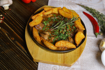 In a frying pan, fried potatoes frame pieces of meat stewed in sauce, cutlery, red bell peppers, champignons, sauce, hot peppers, spices, rosemary branch are on the table. 