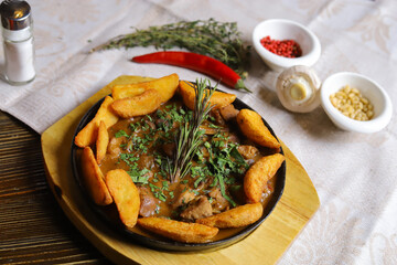 In a frying pan, fried potatoes frame pieces of meat stewed in sauce, cutlery, red bell peppers, champignons, sauce, hot peppers, spices, rosemary branch are on the table. 