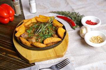 In a frying pan, fried potatoes frame pieces of meat stewed in sauce, cutlery, red bell peppers, champignons, sauce, hot peppers, spices, rosemary branch are on the table. 