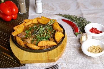 In a frying pan, fried potatoes frame pieces of meat stewed in sauce, cutlery, red bell peppers, champignons, sauce, hot peppers, spices, rosemary branch are on the table. 