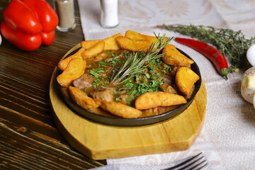 In a frying pan, fried potatoes frame pieces of meat stewed in sauce, cutlery, red bell peppers, champignons, sauce, hot peppers, spices, rosemary branch are on the table. 