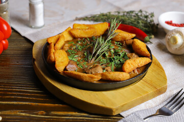 In a frying pan, fried potatoes frame pieces of meat stewed in sauce, cutlery, red bell peppers, champignons, sauce, hot peppers, spices, rosemary branch are on the table. 