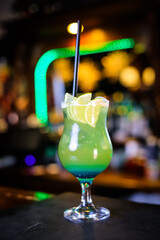 A cocktail in a tall green glass, lemon wedges and a black straw. Filming of the bar menu against the background of bright lights in bokeh.