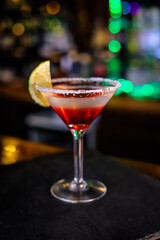 In a funnel-shaped glass on a long stem, a two-tone cocktail, dark red below, translucent white above, a lime slice on the edge of the glass. Filming of the bar menu against the background