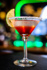 In a funnel-shaped glass on a long stem, a two-tone cocktail, dark red below, translucent white above, a lime slice on the edge of the glass. Filming of the bar menu against the background