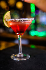 In a funnel-shaped glass on a long stem, a two-tone cocktail, dark red below, translucent white above, a lime slice on the edge of the glass. Filming of the bar menu against the background