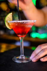 In a funnel-shaped glass on a long stem, a two-tone cocktail, dark red below, translucent white above, a lime slice on the edge of the glass. Filming of the bar menu against the background