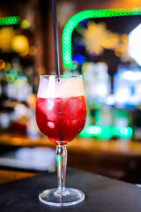 In a tall glass, an opaque, blood-red cocktail with high white foam on top and a black straw. 