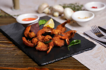 Shrimp is served with sauce. Tempura king prawns, sliced lemon, green basil leaves are served on a black stone stand. Cutlery, hot pepper pods, spices in bowls, white mushrooms.