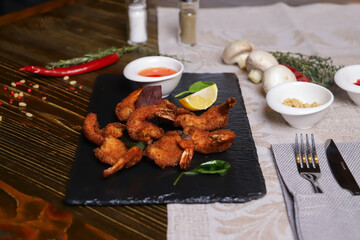 Shrimp is served with sauce. Tempura king prawns, sliced lemon, green basil leaves are served on a black stone stand. Cutlery, hot pepper pods, spices in bowls, white mushrooms.